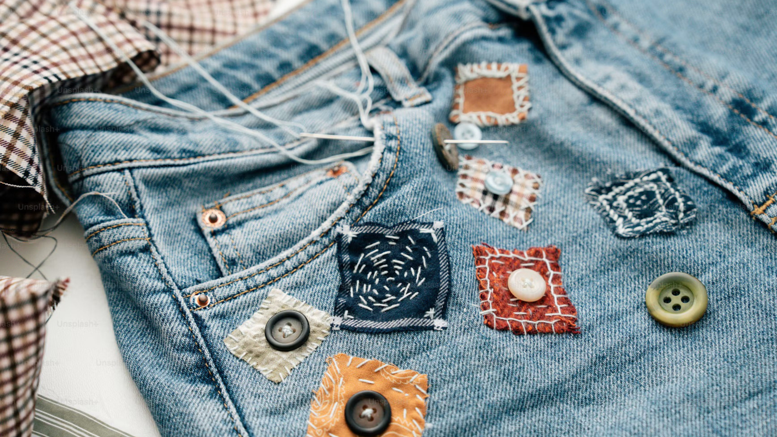 Dettaglio di jeans in denim riparati con toppe di stoffa colorate, bottoni e cuciture a vista di rammendo visibile.